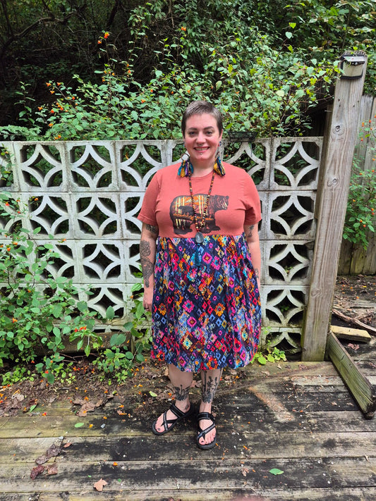 Person wearing a colorful skirt and graphic t-shirt standing on a wooden deck with greenery in the background